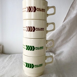 May include: Stack of four white ceramic mugs with brown and green logos. Each mug has the text "Pullman Incorporated" and a triple arrow design. The arrows are brown on the top two mugs and green on the bottom two mugs.