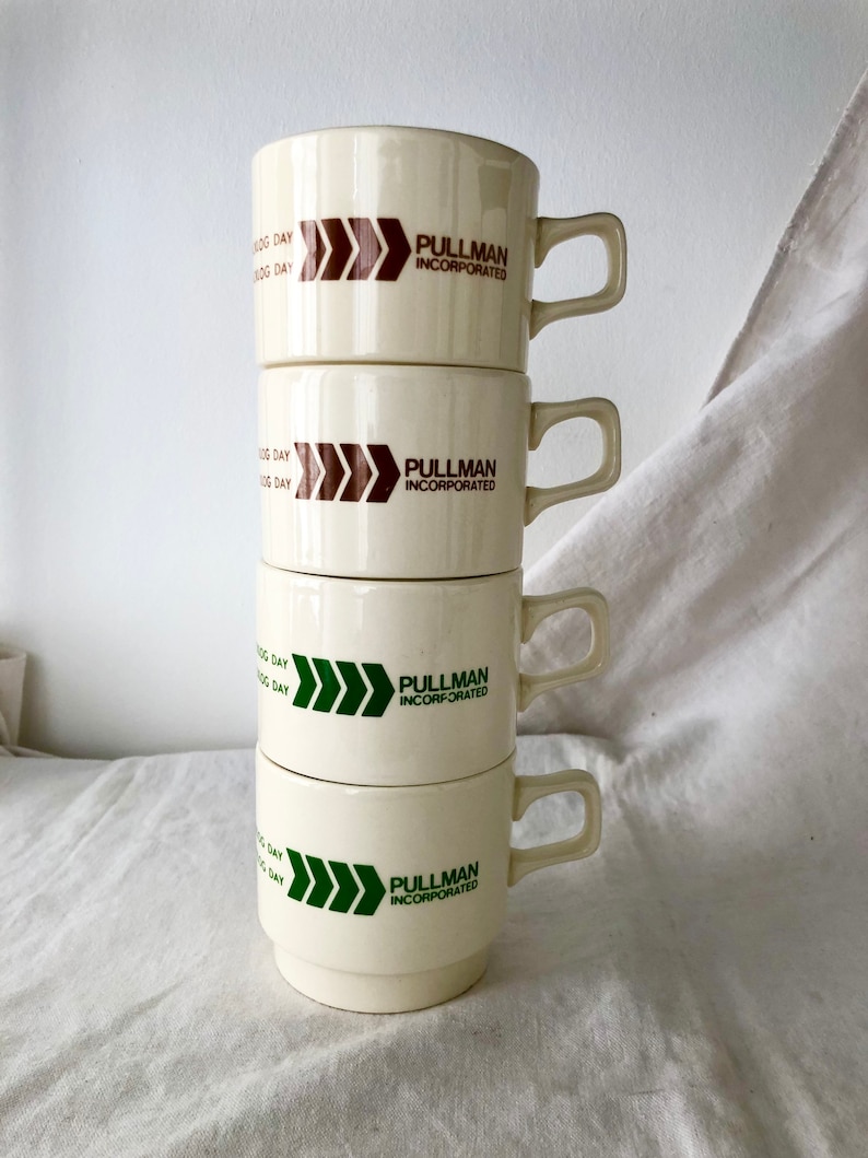May include: Stack of four white ceramic mugs with brown and green logos. Each mug has the text "Pullman Incorporated" and a triple arrow design. The arrows are brown on the top two mugs and green on the bottom two mugs.