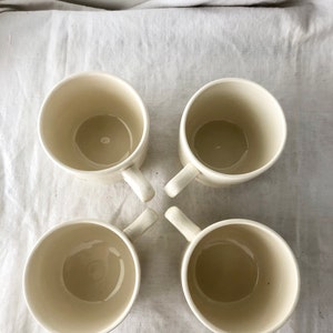 May include: Four white ceramic mugs with handles. The mugs are empty and arranged in a cross pattern on a white fabric surface.