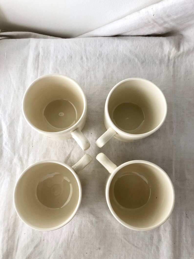 May include: Four white ceramic mugs with handles. The mugs are empty and arranged in a cross pattern on a white fabric surface.