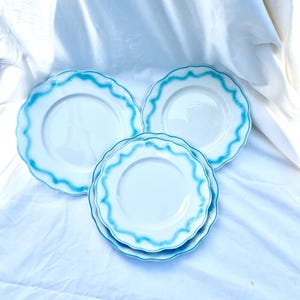 May include: A collection of white ceramic plates with a wavy, turquoise blue trim. The plates are arranged on a white cloth, showcasing their clean design and decorative edge. The set includes both dinner and smaller plates.