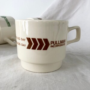 May include: White ceramic coffee mug with a brown logo that says "Pullman Incorporated" and a brown chevron design. The mug has a brown "K-Log Day" inscription.