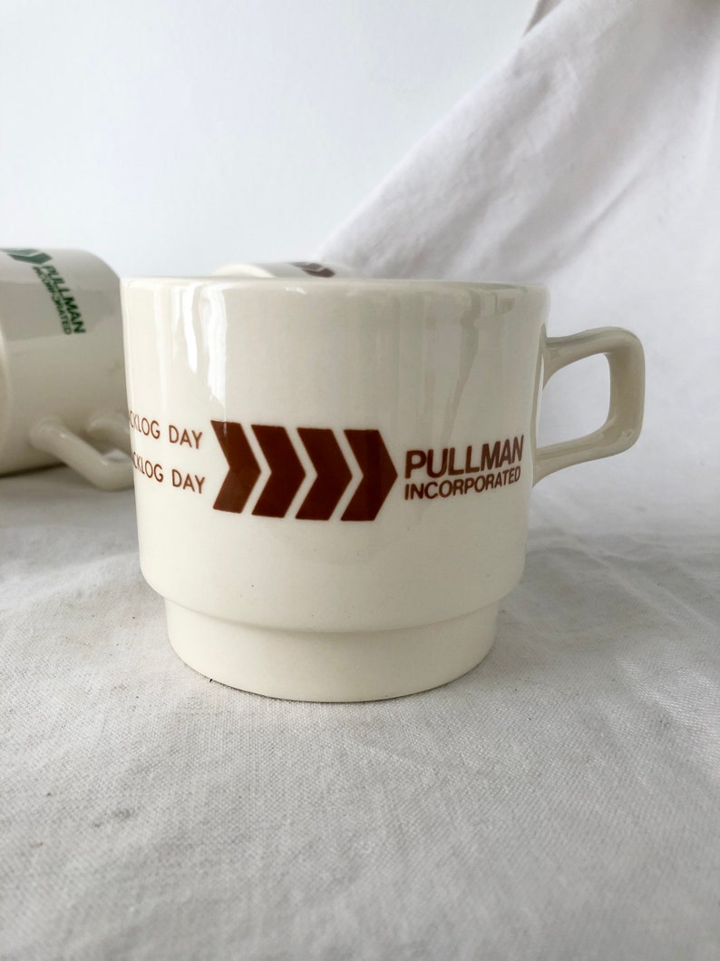 May include: White ceramic coffee mug with a brown logo that says "Pullman Incorporated" and a brown chevron design. The mug has a brown "K-Log Day" inscription.