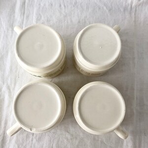 May include: Four white ceramic mugs with handles. The mugs are empty and sitting on a white fabric surface.