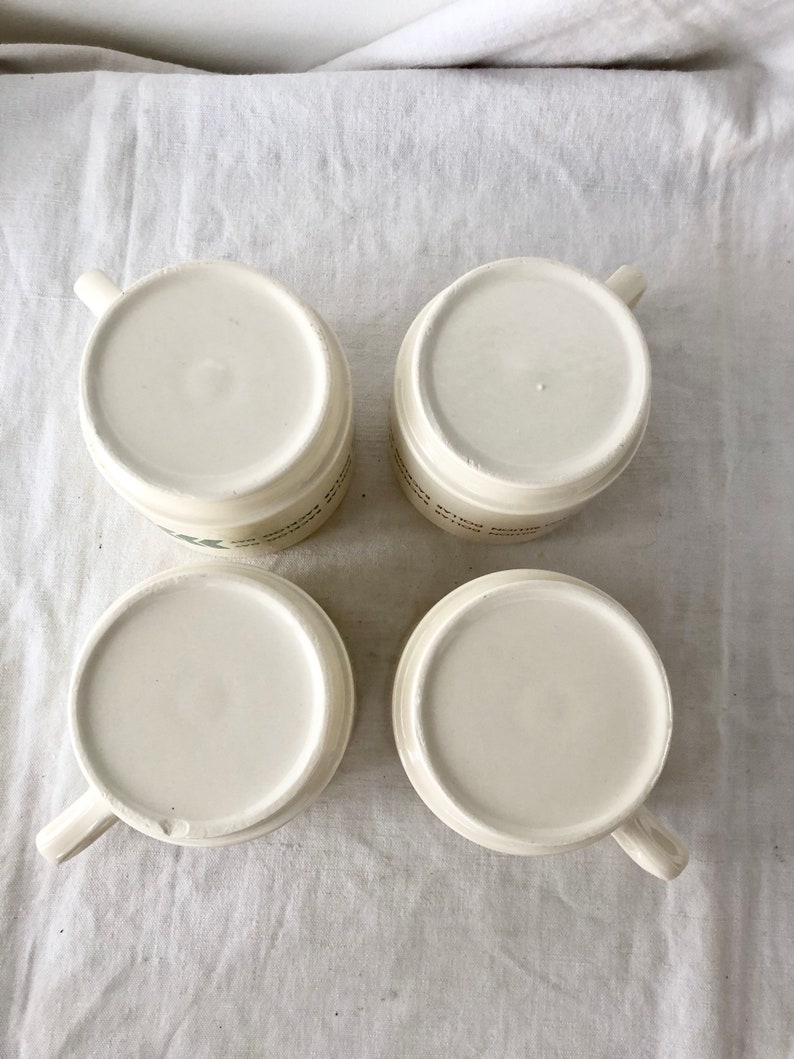 May include: Four white ceramic mugs with handles. The mugs are empty and sitting on a white fabric surface.