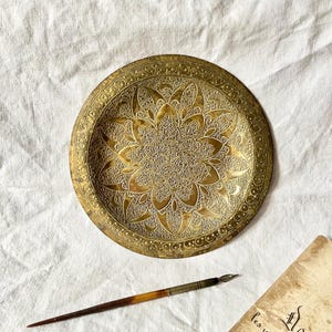 May include: A detailed, antique brass plate with an intricate floral design in the center. A vintage dip pen and a piece of aged paper with script are also present, suggesting a historical or artistic theme.