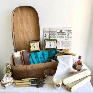 May include: A wooden box with a hinged lid, filled with various items, including two vintage alarm clocks, a glass, a small white porcelain egg cup, a small white porcelain box, a white cloth napkin, two white brushes, a bottle of liquor, and a set of silver forks with ivory handles. The box is sitting on a white tablecloth.