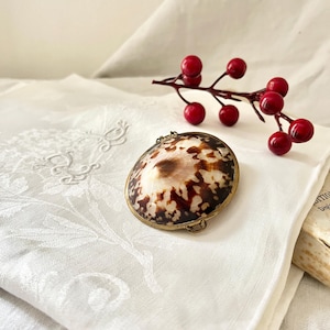 Puede incluir: Una concha redonda, marrón y blanca con un marco y un cierre de latón. La concha está sobre un mantel de lino blanco con un patrón floral.