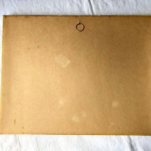 May include: The back of a framed picture with a gold frame. The back of the frame is made of brown cardboard and has a small metal ring in the upper centre. There is some writing in the top right corner of the back of the frame.