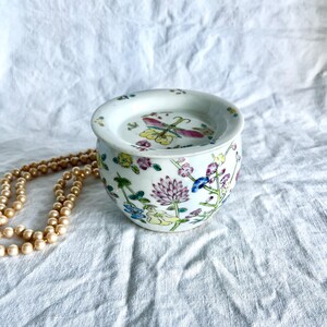 Pode incluir: Um pequeno frasco redondo de porcelana branca com tampa a condizer, decorado com motivos florais coloridos pintados à mão e uma libélula. O frasco tem um acabamento liso e brilhante e é acompanhado por um colar de pérolas claras.
