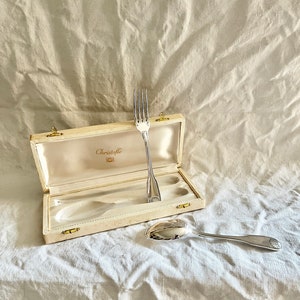Peut inclure: Une fourchette et une cuillère en argent dans un coffret blanc avec des accents dorés. Le coffret est marqué "Christofle".