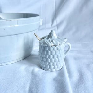 May include: A small, white ceramic sugar bowl with a lid and a small handle. The bowl has a textured, honeycomb-like pattern. A small wooden spoon rests inside the bowl. A larger white ceramic bowl is partially visible in the background.