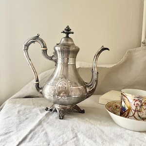 Peut inclure: Une théière en argent avec un design orné et un motif floral. La théière a un couvercle avec un bouton et un bec verseur. La théière est posée sur une nappe blanche.
