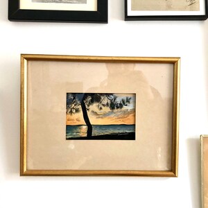 May include: A framed watercolour painting of a silhouette of a tree on a beach at sunset. The painting is in a gold frame and is hanging on a white wall.