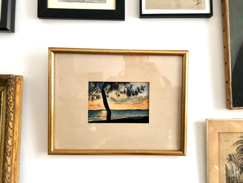 May include: A framed watercolour painting of a silhouette of a tree on a beach at sunset. The painting is in a gold frame and is hanging on a white wall.