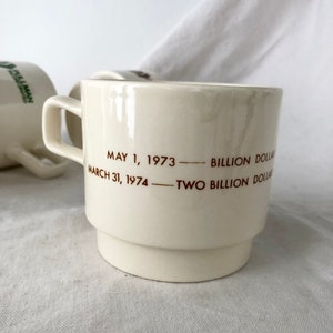 May include: White ceramic coffee mug with brown text that reads "May 1, 1973 - Billion Dollar - March 31, 1974 - Two Billion Dollar"