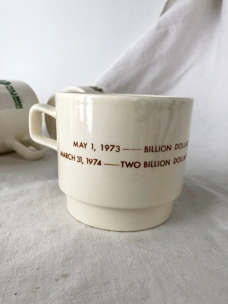 May include: White ceramic coffee mug with brown text that reads "May 1, 1973 - Billion Dollar - March 31, 1974 - Two Billion Dollar"