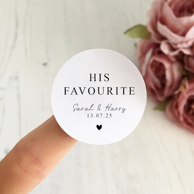 May include: A white, circular sticker with the text "HIS FAVOURITE" in a black, elegant font. Below, it reads "Sarah & Harry" and the date "13.07.25", with a small black heart at the bottom. The sticker is held by a finger.