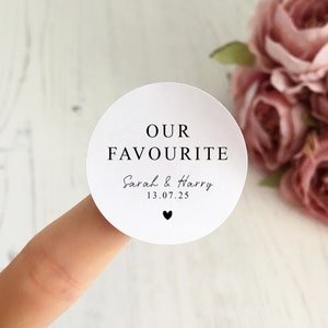 May include: A white, circular sticker with the words "OUR FAVOURITE" in a bold, black font. Below, in a script font, are the names "Sarah & Harry" and the date "13.07.25", with a small black heart beneath.