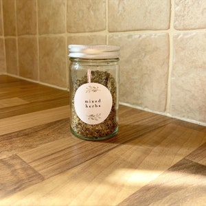 K&ouml;nnte beinhalten: Ein Glasgef&auml;&szlig; mit silbernem Deckel, gef&uuml;llt mit getrockneten Kr&auml;utern. Das Gef&auml;&szlig; hat ein wei&szlig;es Etikett mit dem Text "mixed herbs" und einem kleinen floralen Design.