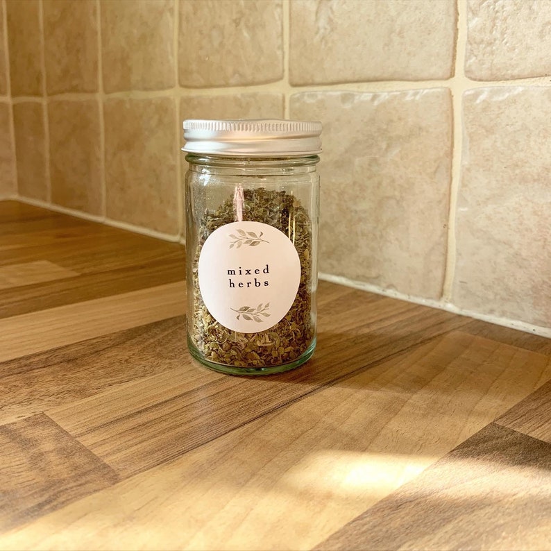 K&ouml;nnte beinhalten: Ein Glasgef&auml;&szlig; mit silbernem Deckel, gef&uuml;llt mit getrockneten Kr&auml;utern. Das Gef&auml;&szlig; hat ein wei&szlig;es Etikett mit dem Text "mixed herbs" und einem kleinen floralen Design.