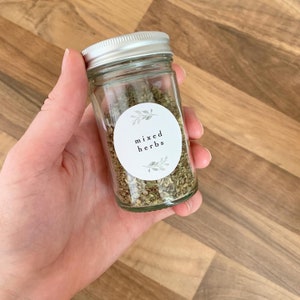 K&ouml;nnte beinhalten: Ein Glasgef&auml;&szlig; mit silbernem Deckel, gef&uuml;llt mit getrockneten Kr&auml;utern. Das Gef&auml;&szlig; hat ein wei&szlig;es Etikett mit gr&uuml;nen Bl&auml;ttern und dem Text "mixed herbs".