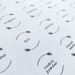 Op de afbeelding: Een set witte ronde labels met zwarte tekst en een eenvoudige tekening van een tarwehalm. De labels bevatten de woorden: oregano, rozemarijn, peterselie, dille, venkelzaad, komijnpoeder.
