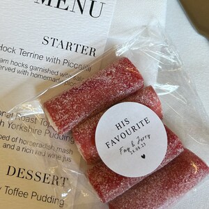 May include: A clear plastic bag containing several red, sugar-coated candies. A white circular sticker on the bag reads "HIS FAVOURITE Fay & Jerry 24.08.23". A menu is visible in the background.