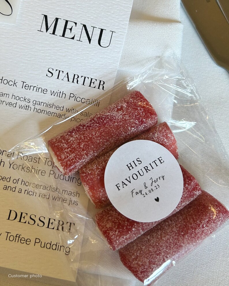 May include: A clear plastic bag containing several red, sugar-coated candies. A white circular sticker on the bag reads "HIS FAVOURITE Fay & Jerry 24.08.23". A menu is visible in the background.