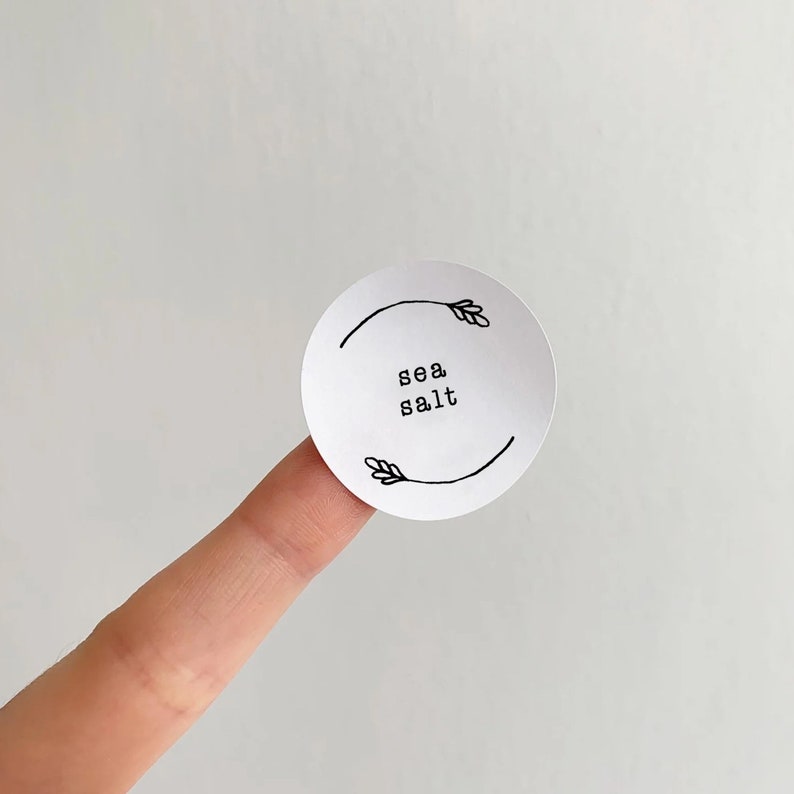 Op de afbeelding: Ronde witte sticker met zwarte tekst en een eenvoudige lijntekening van een tak met bladeren aan elke kant. De tekst luidt "sea salt".