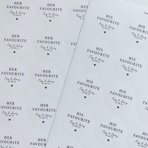 May include: Sheets of white, circular stickers with black text. The stickers read "HER FAVOURITE" and "HIS FAVOURITE" with a date and a heart symbol. The stickers are for a wedding or special event.