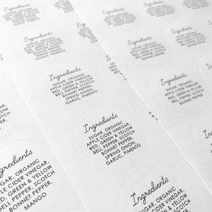 May include: Close-up of several white round stickers with black text. The text reads "Ingredients: Sugar, Organic Apple Cider Vinegar, Red, Green & Yellow Bell Pepper, Scotch Bonnet Pepper, Spring Onion, Garlic, Pimento".