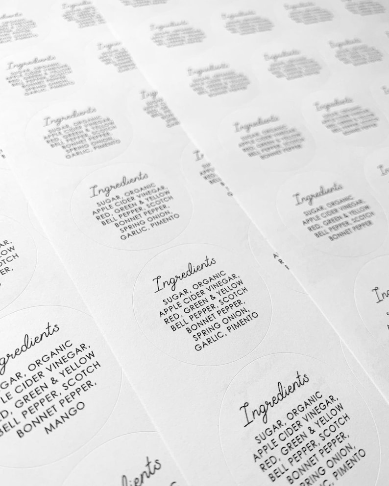 May include: Close-up of several white round stickers with black text. The text reads "Ingredients: Sugar, Organic Apple Cider Vinegar, Red, Green & Yellow Bell Pepper, Scotch Bonnet Pepper, Spring Onion, Garlic, Pimento".