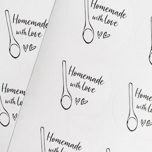 K&ouml;nnte beinhalten: Wei&szlig;e runde Aufkleber mit schwarzem Text und einer Zeichnung eines Holzl&ouml;ffels. Der Text lautet "Hausgemacht mit Liebe" und enth&auml;lt zwei kleine Herzen.