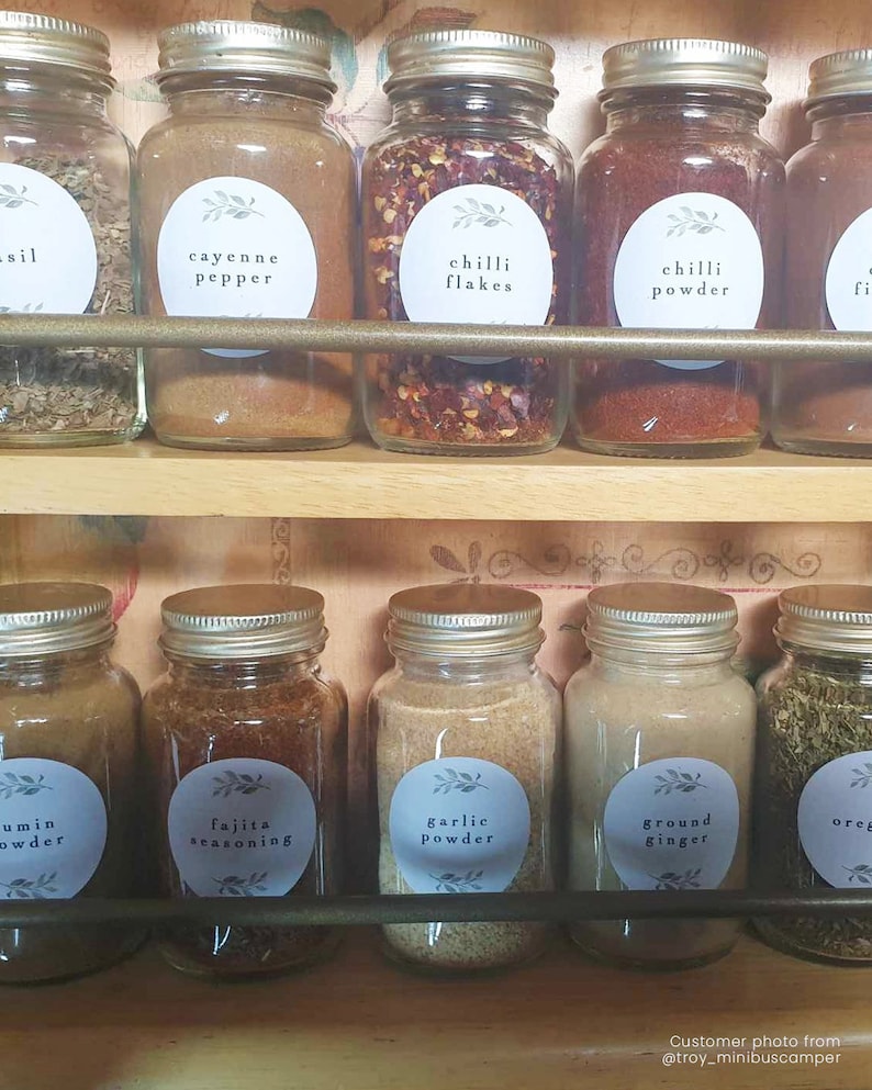 K&ouml;nnte beinhalten: Ein Satz Glas-Gew&uuml;rzgl&auml;ser mit Etiketten f&uuml;r verschiedene Gew&uuml;rze, darunter Cayennepfeffer, Chiliflocken, Chilipulver, Kreuzk&uuml;mmelpulver, Fajita-Gew&uuml;rz, Knoblauchpulver, gemahlener Ingwer und Oregano. Die Gl&auml;ser sind auf einem Holzregal angeordnet.