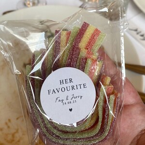 May include: A clear cellophane bag filled with colorful, layered candy. The candy is a mix of red, yellow, and green. A white circular sticker on the bag reads "HER FAVOURITE" with the names "Fay & Jerry" and the date "24.08.23".