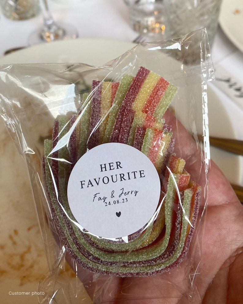 May include: A clear cellophane bag filled with colorful, layered candy. The candy is a mix of red, yellow, and green. A white circular sticker on the bag reads "HER FAVOURITE" with the names "Fay & Jerry" and the date "24.08.23".