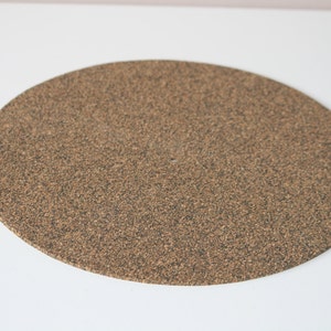 May include: A round, brown cork turntable slipmat. The slipmat is made of a textured cork material.