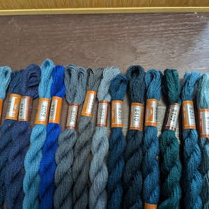 Könnte beinhalten: Eine Sammlung von 16 Knäuel Stickgarn in Blautönen und Grüntönen. Das Garn ist in einem Farbverlauf von Hellblau bis Dunkelgrün angeordnet. Jedes Knäuel ist mit einer Nummer gekennzeichnet.