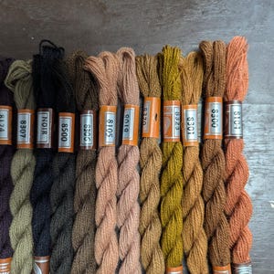 May include: A close-up of 14 spools of embroidery floss in various shades of brown, beige, and orange. The spools are labelled with numbers and the words "Blanc", "Noir", and "8500".