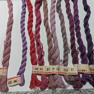 May include: A collection of embroidery floss in various shades of pink, purple, and red. The floss is twisted into ropes and arranged on a white surface. Each color has a small tag with numbers and the word "ISTERGARN".