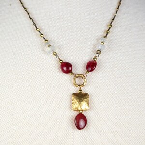 Könnte beinhalten: Handgefertigte Halskette mit goldfarbener Kette, kleinen goldenen Perlen und roten und weißen Edelsteinakzenten. Der Anhänger hat ein gehämmertes Quadrat und einen tropfenförmigen roten Edelstein.