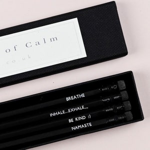 May include: A black box with a white label that says "Moments of Calm" and a website address. The box contains four black pencils with white text that reads "Breathe", "Inhale...Exhale...", "Be Kind :)", and "Namaste".