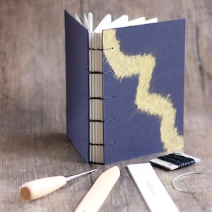 May include: A navy blue book with a gold zigzag design on the cover. The book is bound with a coptic stitch and has a spine made of natural paper. The image also shows tools for bookbinding, including a bone folder, an awl, and a needle.