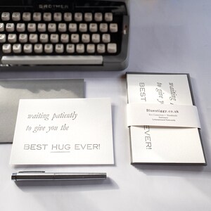 May include: Two white greeting cards with gray borders. The top card reads "waiting impatiently to give you the BEST HUG EVER!" The bottom card reads "waiting impatiently to give you the BEST EVER!" The cards are on a white surface with a silver pen and a vintage typewriter.
