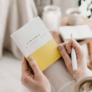 Puede incluir: Una tarjeta blanca con papel amarillo en la mitad inferior. La tarjeta tiene el texto "All the reasons why I love you" escrito en tipografía de máquina de escribir.