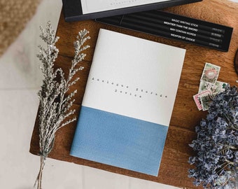 May include: A blue and white notebook with the text "Analogue Storage Device" printed on the front. The notebook is sitting on a wooden surface with a box of pencils and some stamps.