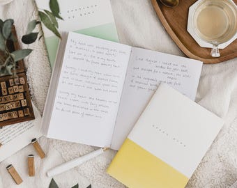May include: A white and yellow notebook with the text "Green Book" on the cover. The notebook is open to a page with handwritten text. The text includes the words "My heart was wondering my soul" and "Lost August I looked out the window by your bed". The notebook is on a white fuzzy surface with a glass teapot, a cup of tea, and a wooden tray.