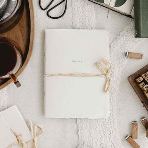 May include: A white journal tied with twine and a cream-coloured ribbon, with the word "Herke" printed on the cover. The journal is on a textured white surface, with a pair of black scissors, a cup of tea, and a book in the background.