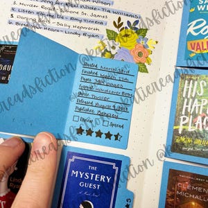 May include: A close-up of a blue book journal with a blue bookmark. The bookmark has a handwritten review of a book titled "The Mystery Guest". The review includes the date started, date finished, number of pages, format, genre, release date, publisher, and a 4-star rating.